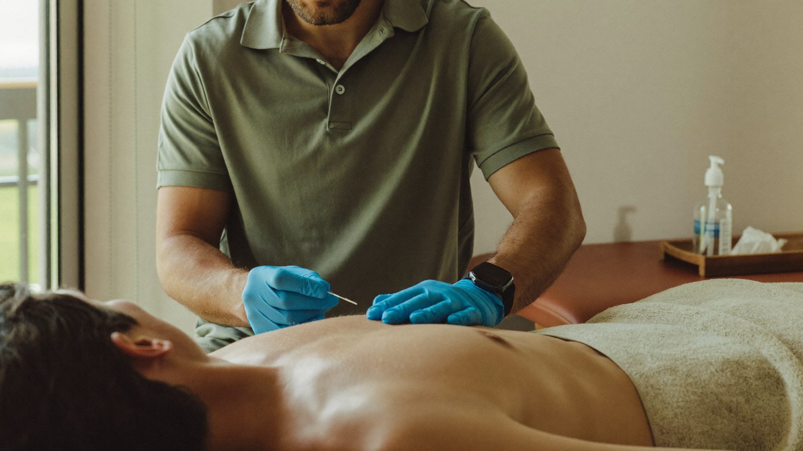 Fisioterapeuta con guantes aplicando punción seca en el trapecio de un paciente tumbado boca abajo en la camilla